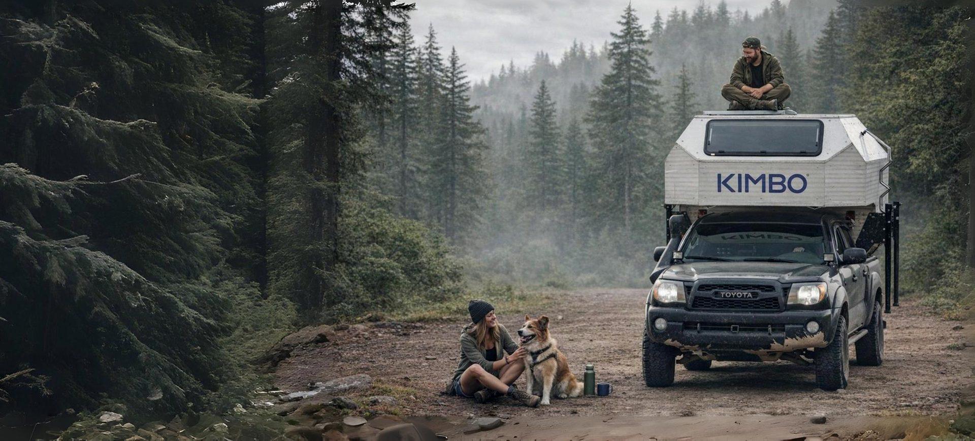 Two Kimbo owners camped in a misty Pacific Northwest forest — one perched on the cabover roof of a white Kimbo hard-side aluminum truck bed camper, another seated by a small fire below with a dog, towering Douglas firs in every direction.