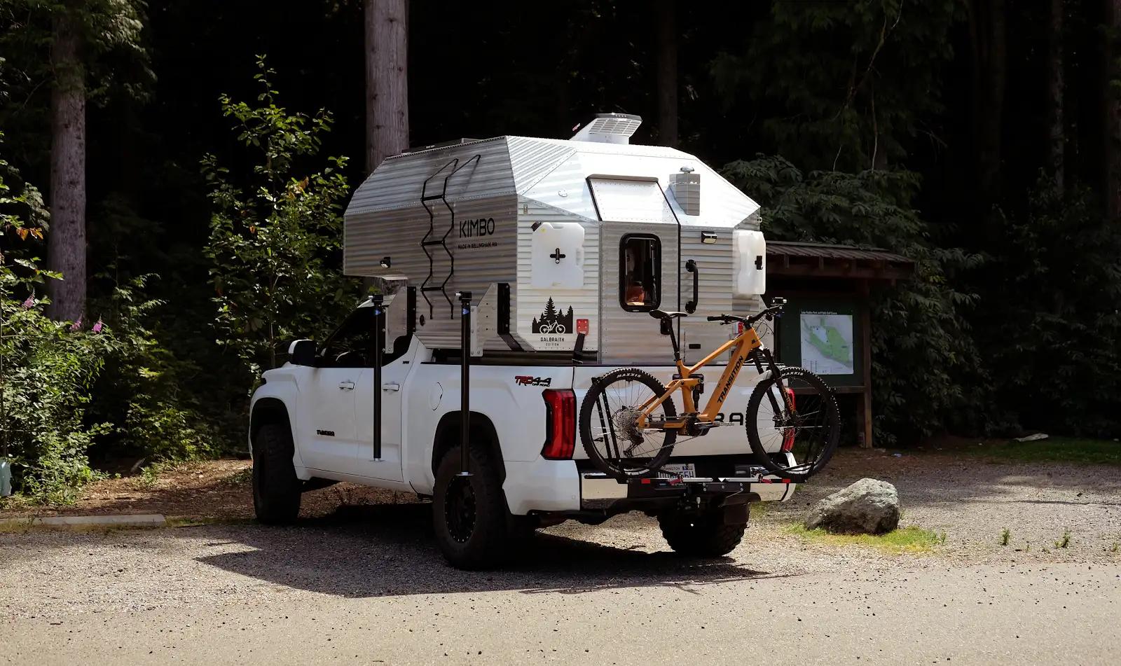 Kimbo 6 Galbraith Edition camper on a white Toyota Tundra parked at a forested Pacific Northwest trailhead, with a mountain bike loaded on the rear rack and the custom Galbraith mountain graphics visible on the camper door.