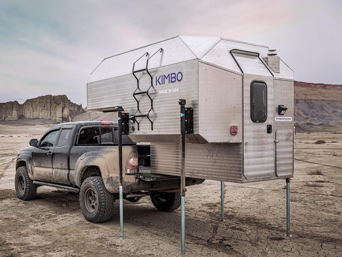 White Toyota Tacoma reverse-loading a Kimbo 6 hard-side aluminum truck bed camper raised on its four corner jacks at a red sandstone overlook in southern Utah.