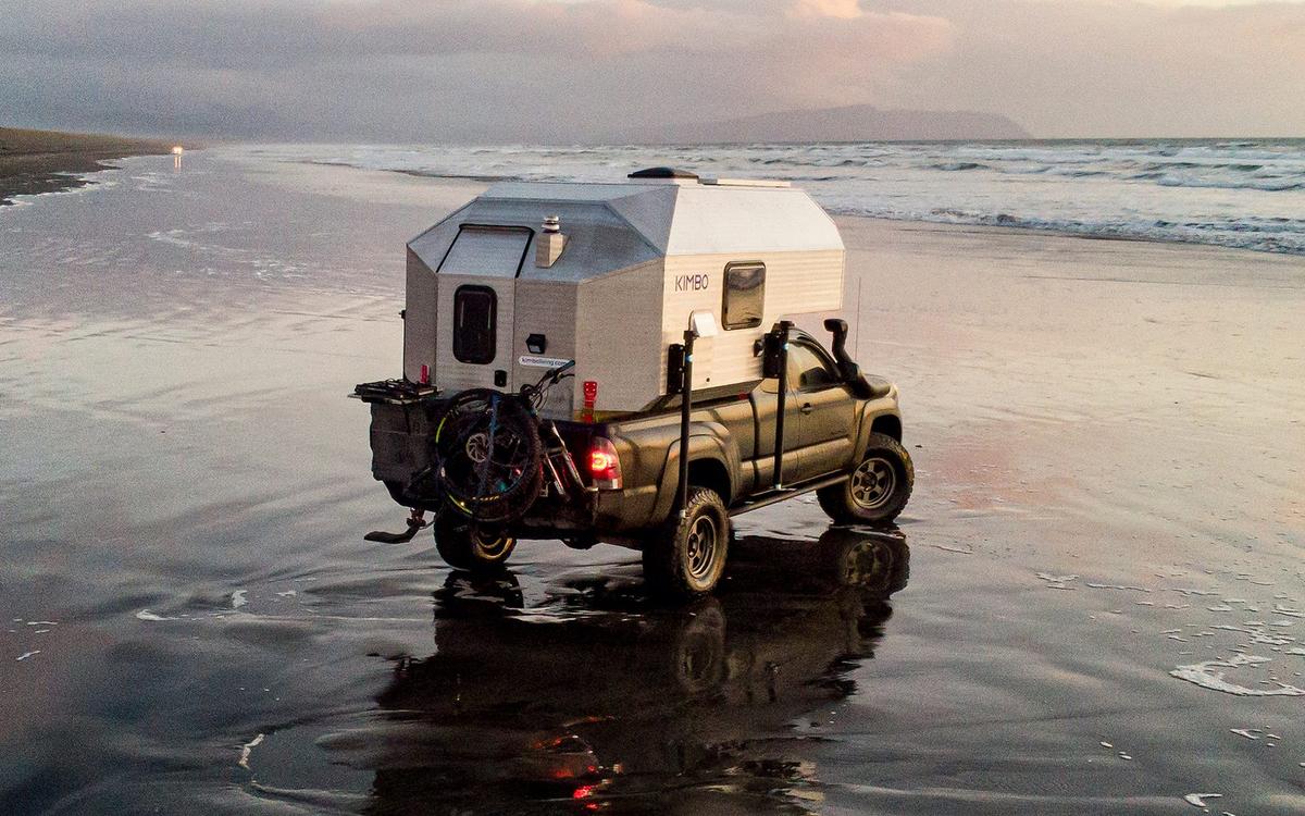 Black Toyota Tacoma with Kimbo 6 hard-side aluminum truck bed camper driving along a misty Pacific Northwest tideline, the truck reflecting in wet sand with a coastal headland fading into the fog behind.