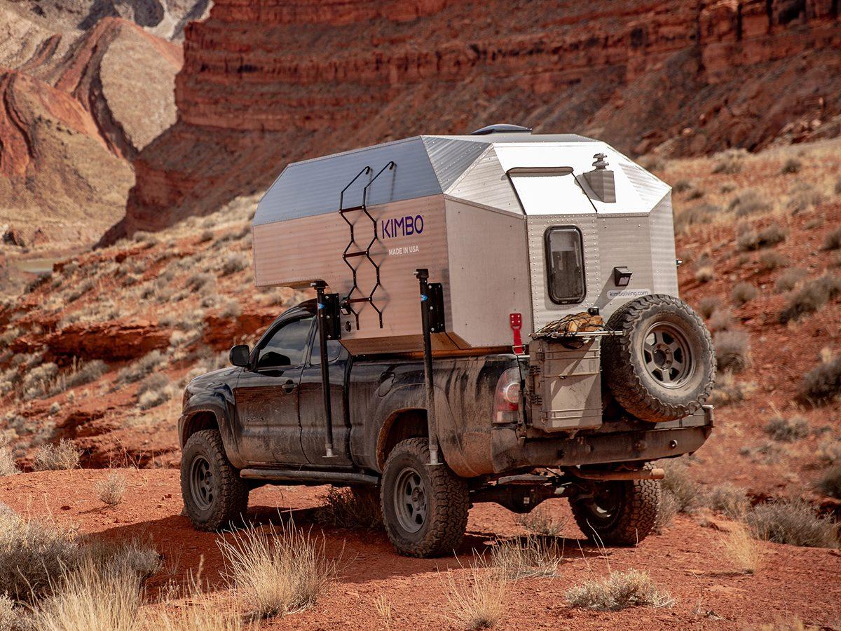 Black Toyota Tacoma with Kimbo 6 hard-side aluminum truck bed camper parked rear-quarter on red dirt at a southern Utah canyon overlook, full-size spare tire mounted on the rear swing-out, sandstone walls behind.