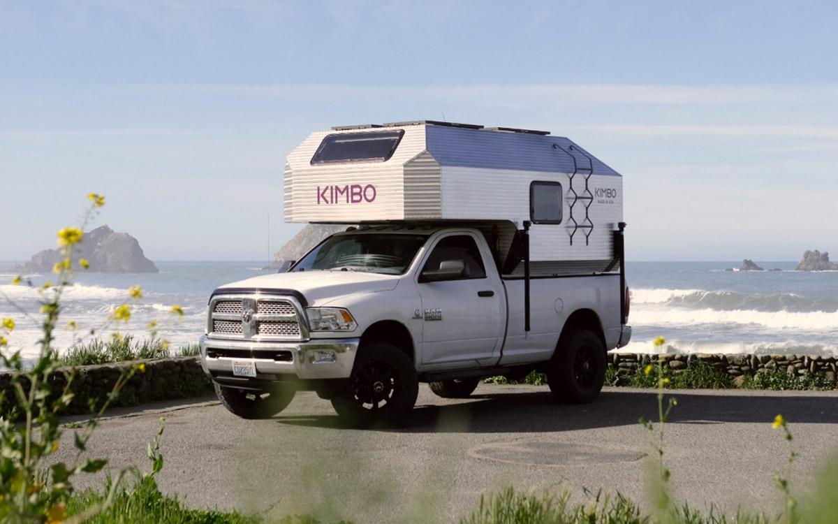 White Dodge Ram 2500 with the Kimbo 8 hand-riveted aluminum truck bed camper installed, photographed at a coastal overlook on the Oregon coast with yellow mustard wildflowers blooming in the foreground, sea stacks and breaking surf in the distance behind the camper.