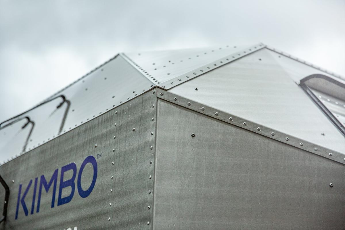 Close-up architectural shot of the hand-riveted aluminum monocoque shell of a Kimbo 6 truck bed camper, KIMBO wordmark on the sidewall, against a moody overcast Pacific Northwest sky.