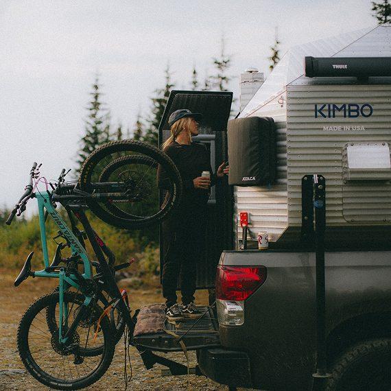 Kimbo owner Amanda standing at the open tailgate of her Kimbo truck bed camper at a Pacific Northwest forest pull-off, with two mountain bikes mounted on the rear rack.