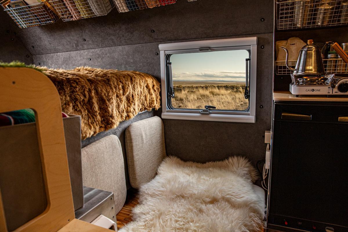 Cabover sleeping nook of a Kimbo 6 hard-side aluminum truck bed camper with a sheepskin throw on the bed, a kettle on the side counter, and an Arctic Tern double-pane window framing a wide prairie horizon.