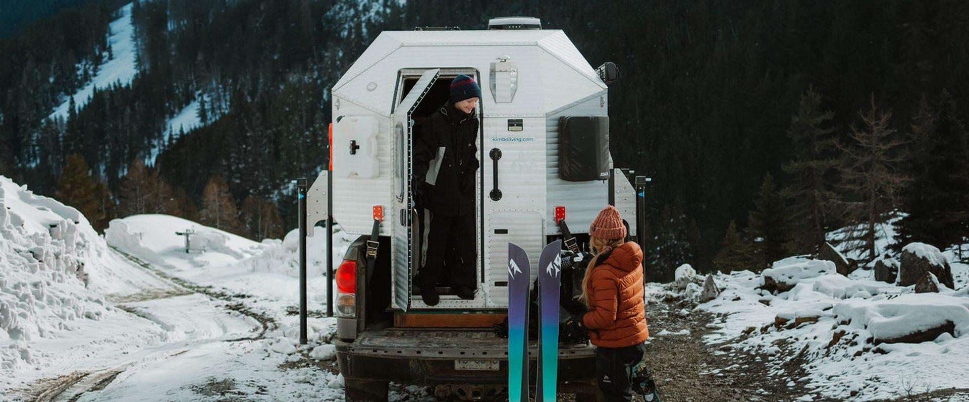 Kimbo owner standing in the open rear doorway of a white Kimbo hard-side aluminum truck bed camper at a snowy Pacific Northwest pull-off, skis racked along the side and a friend in a knit cap waiting outside in the snow.