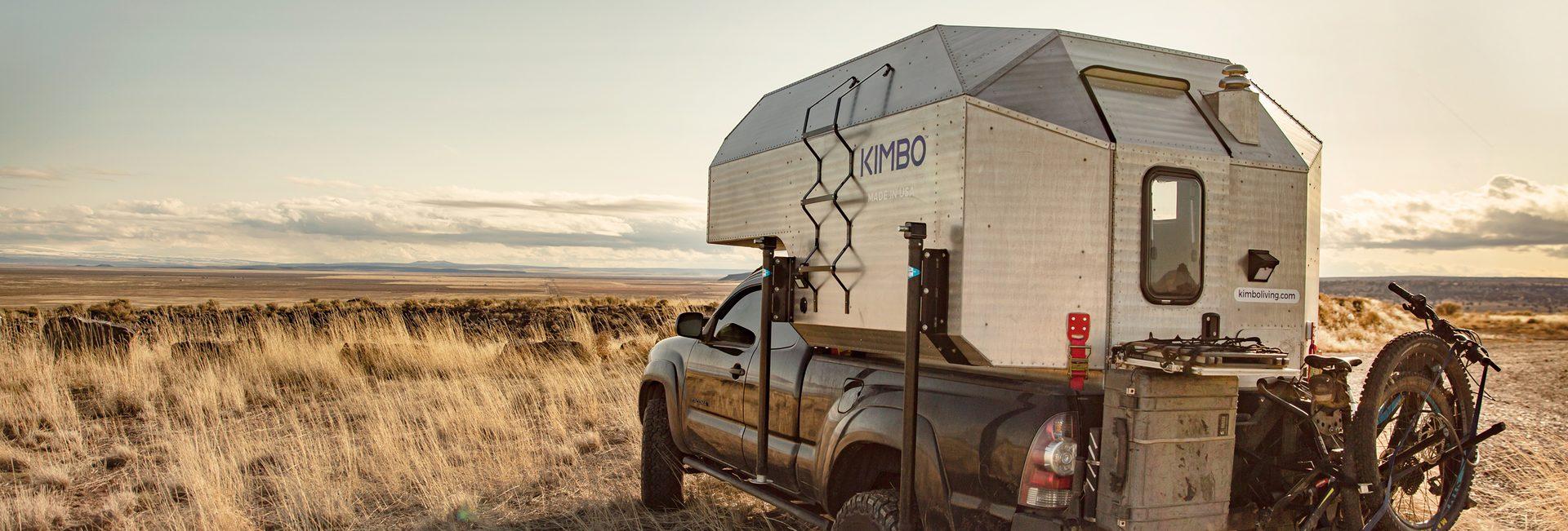 Black Toyota Tacoma with Kimbo 6 hard-side aluminum truck bed camper parked rear-quarter on a sage-prairie pull-off at golden hour, KIMBO wordmark visible, mountain bikes mounted on the rear rack, wide horizon stretching to distant ridges.