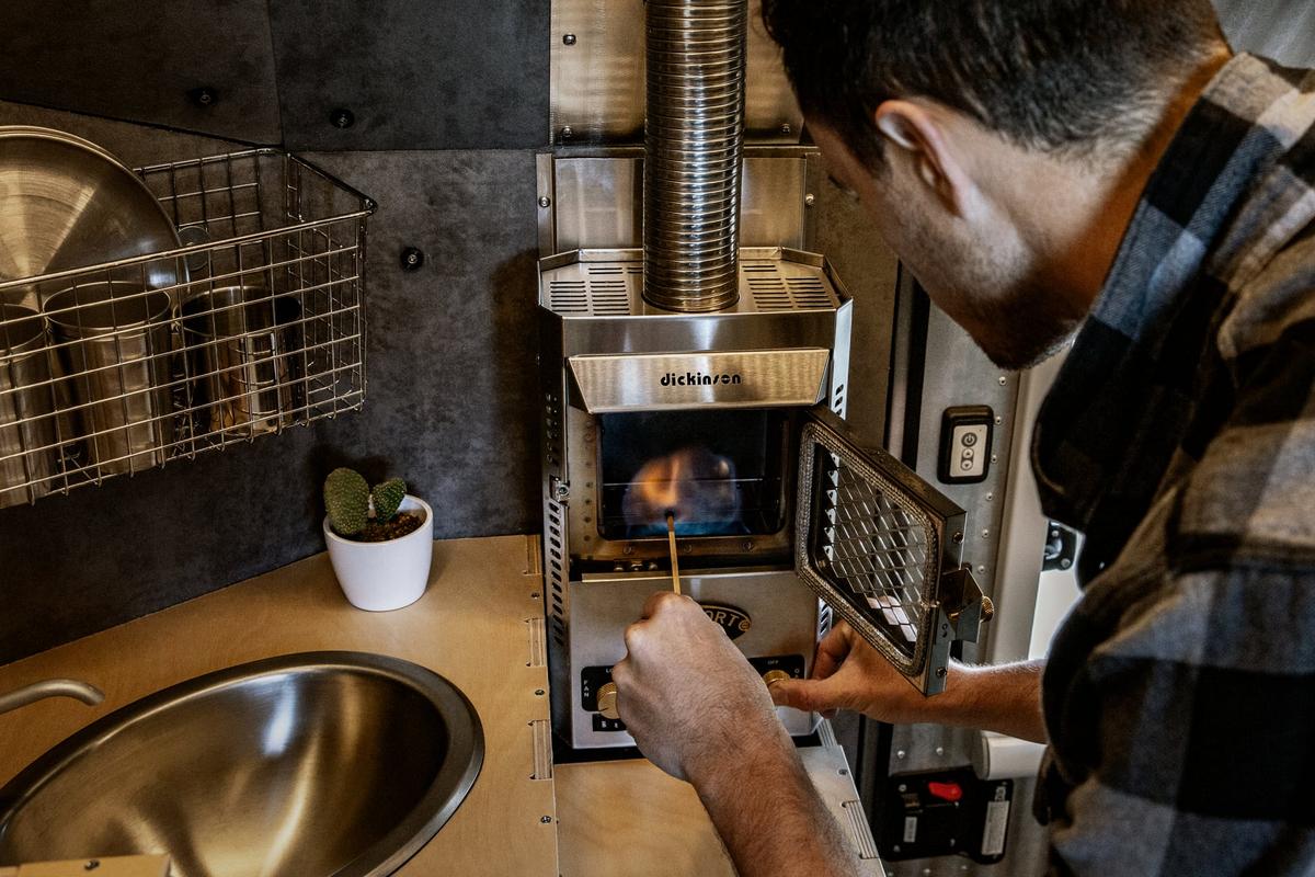 Mark King lighting the Dickinson Marine propane heater installed in a Kimbo 6 hard-side aluminum truck bed camper galley, deep stainless sink and bamboo prep counter visible to the side.