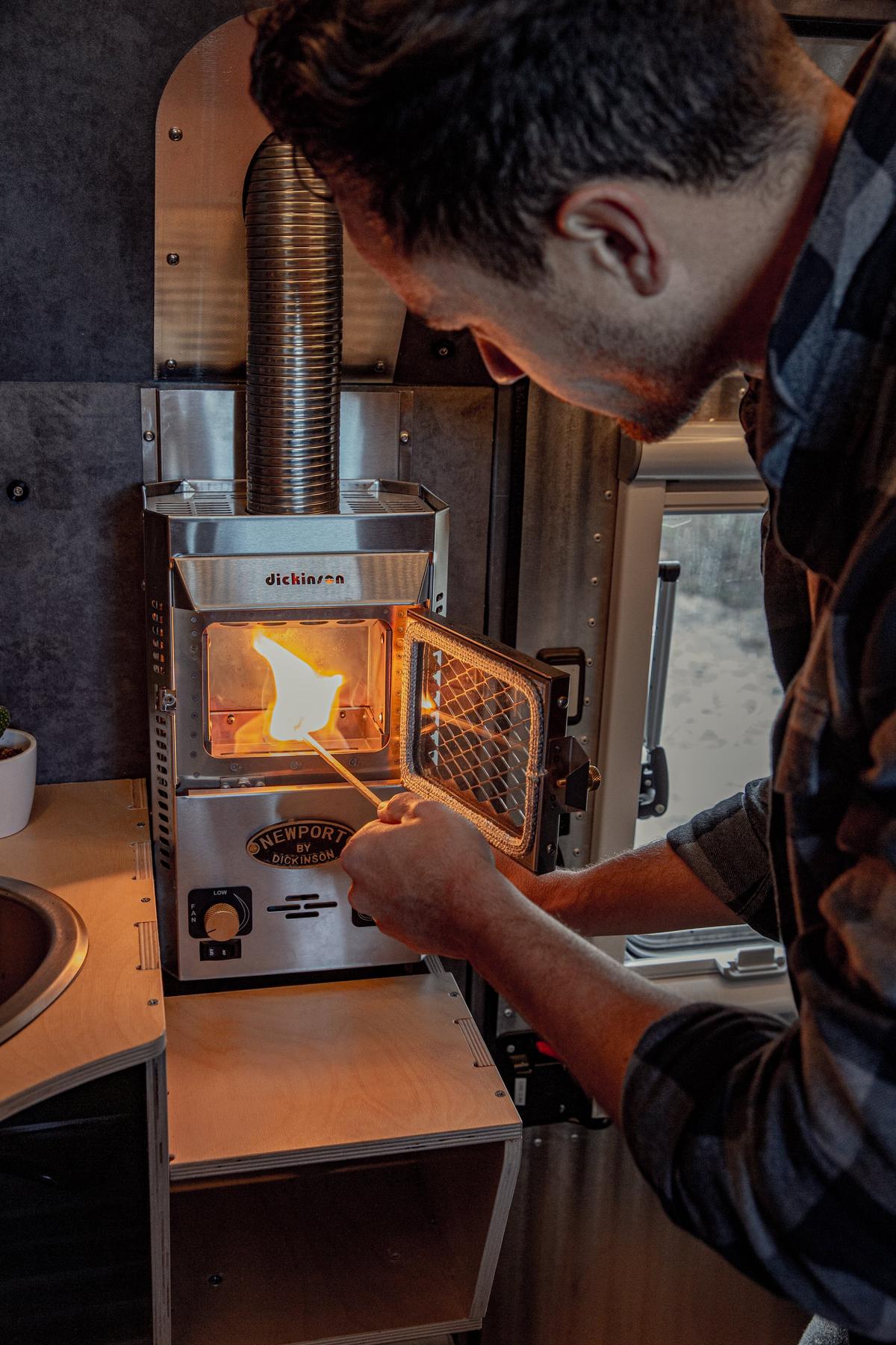 Close-up of the Dickinson Marine propane heater inside a Kimbo 6 hard-side aluminum truck bed camper, an open flame visible through the spark-arrestor door as Mark King reaches in with a long match to light it.