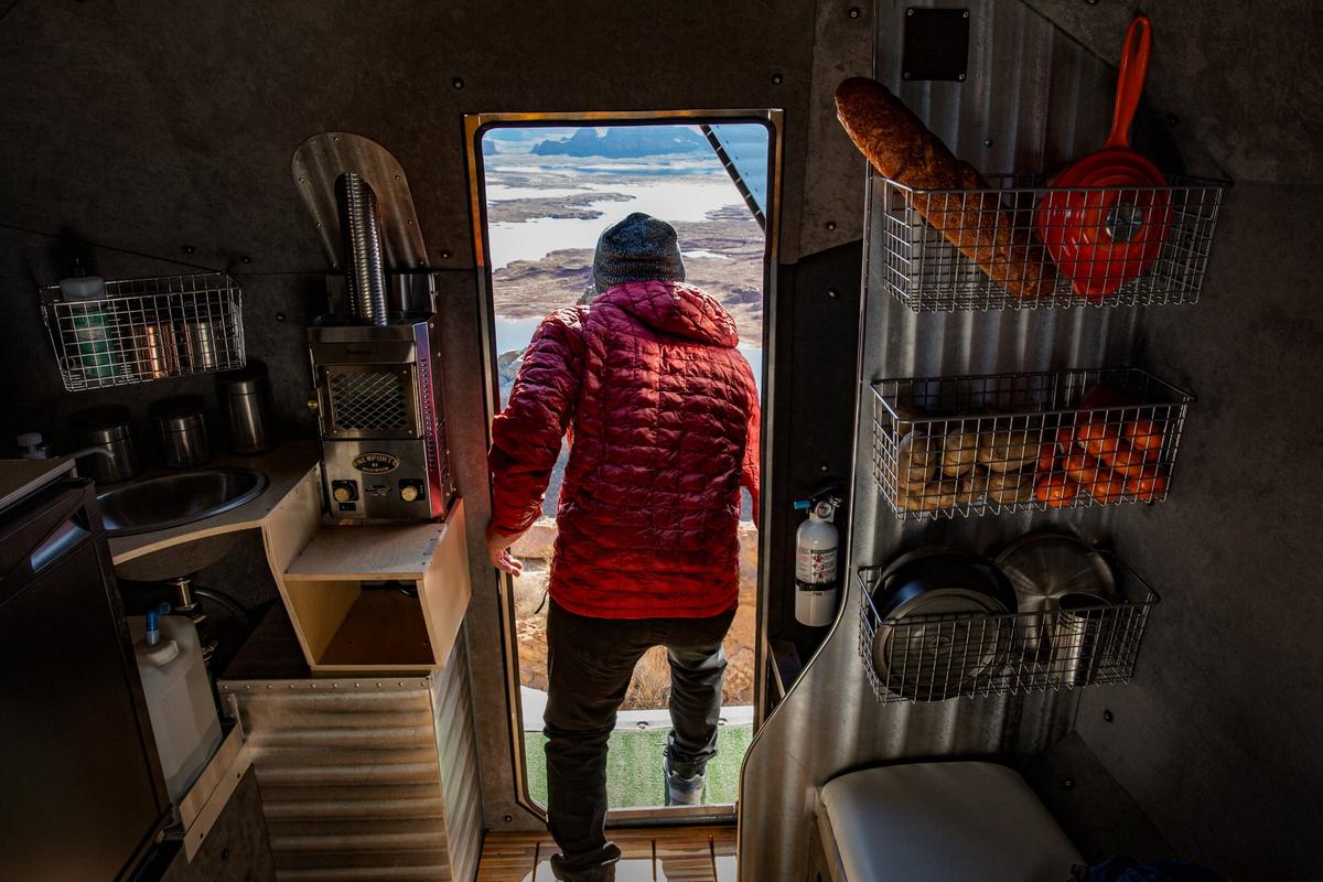 Owner in a red insulated jacket standing in the open doorway of a Kimbo 6 hand-riveted aluminum truck bed camper, looking out at a vast red rock canyon vista with a lake below, wire-basket pantry and stainless sink visible inside.