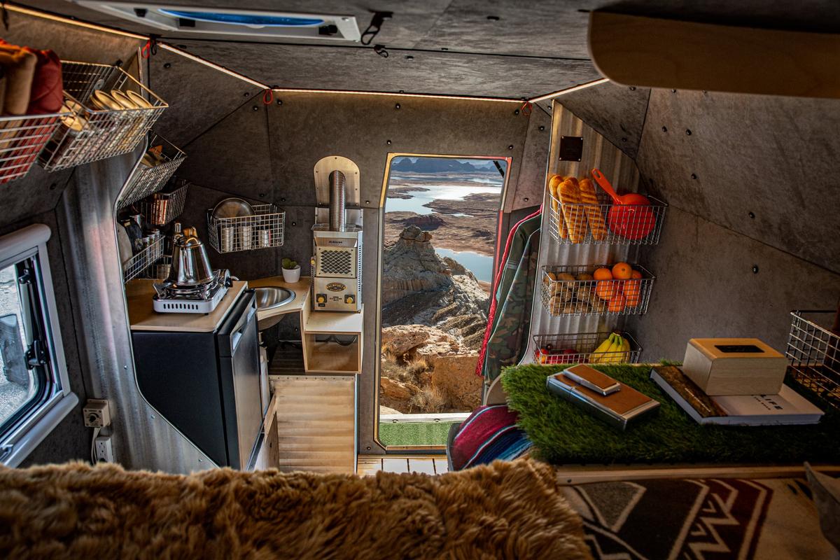 Looking out from inside a Kimbo 6 hard-side aluminum truck bed camper galley — wire-basket pantry full of fruit, stainless sink, induction cooktop, and the open doorway framing a red rock canyon vista with a lake below.