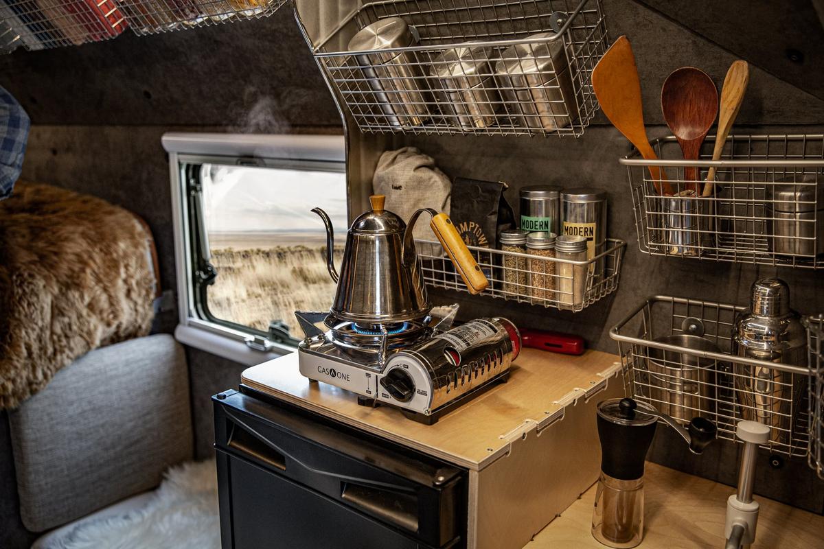 Stainless gooseneck kettle steaming on a portable butane stove inside a Kimbo 6 hard-side aluminum truck bed camper galley, hand-tools and condiments organized in mounted wire-basket pantry, a wide prairie window-view to the side.