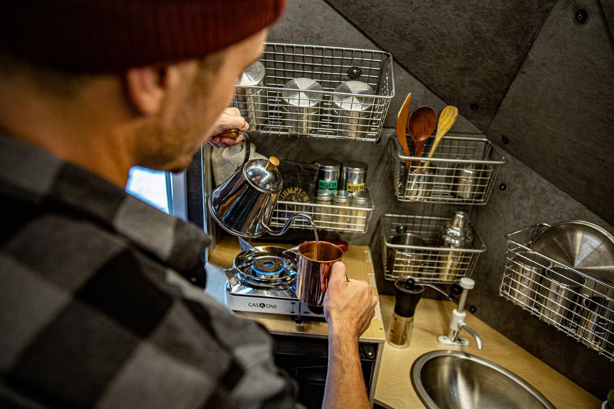 Person in a beanie pouring a pour-over coffee from a stainless gooseneck kettle into a copper mug on the bamboo galley counter of a Kimbo 6 hard-side aluminum truck bed camper, mounted wire-basket pantry stocked with cups and utensils.
