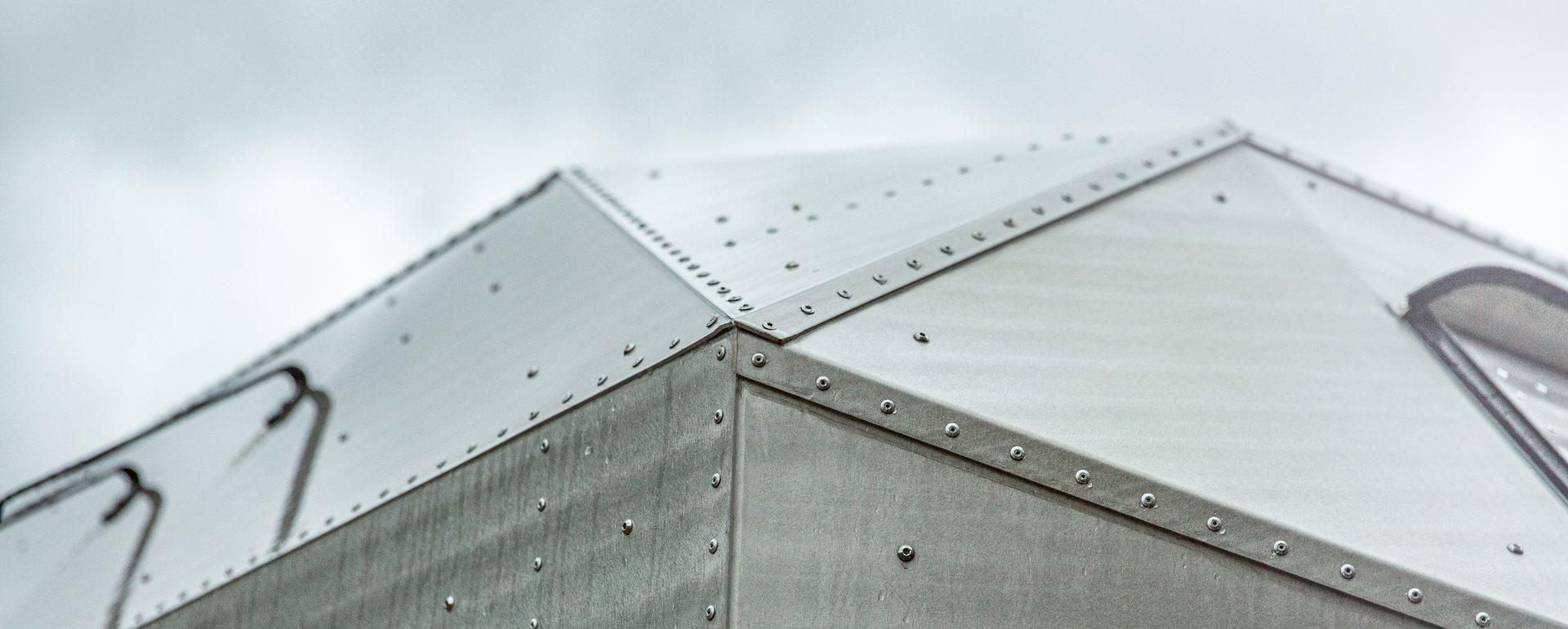 Close-up of the hand-riveted aluminum roof apex of a Kimbo hard-side aluminum truck bed camper, showing the laser-cut 5052 aluminum panels seamed together with solid rivets at 1.5″ spacing and the integrated black side ladder.