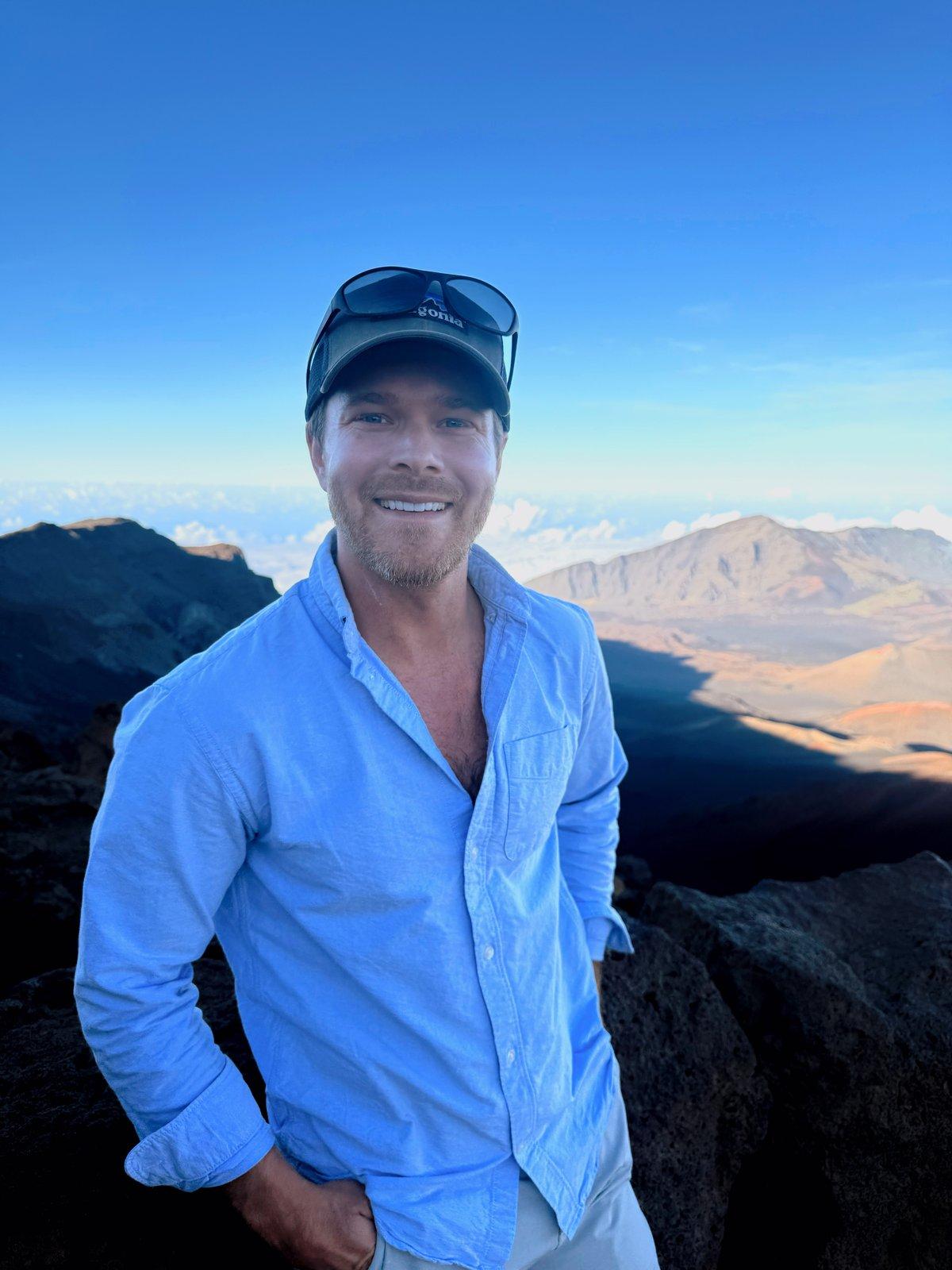 Mark King, founder of Kimbo Living, photographed near the summit at sunrise above the cloud line — wearing a Kimbo cap and a light-blue oxford shirt, with volcanic mountains and a low cloud layer behind him.