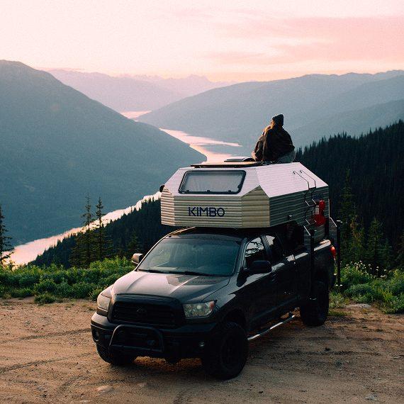Black Toyota Tacoma with Kimbo 6 hard-side aluminum truck bed camper parked on a ridge above a deep alpine valley at sunset, two figures on the cabover roof taking in the view.