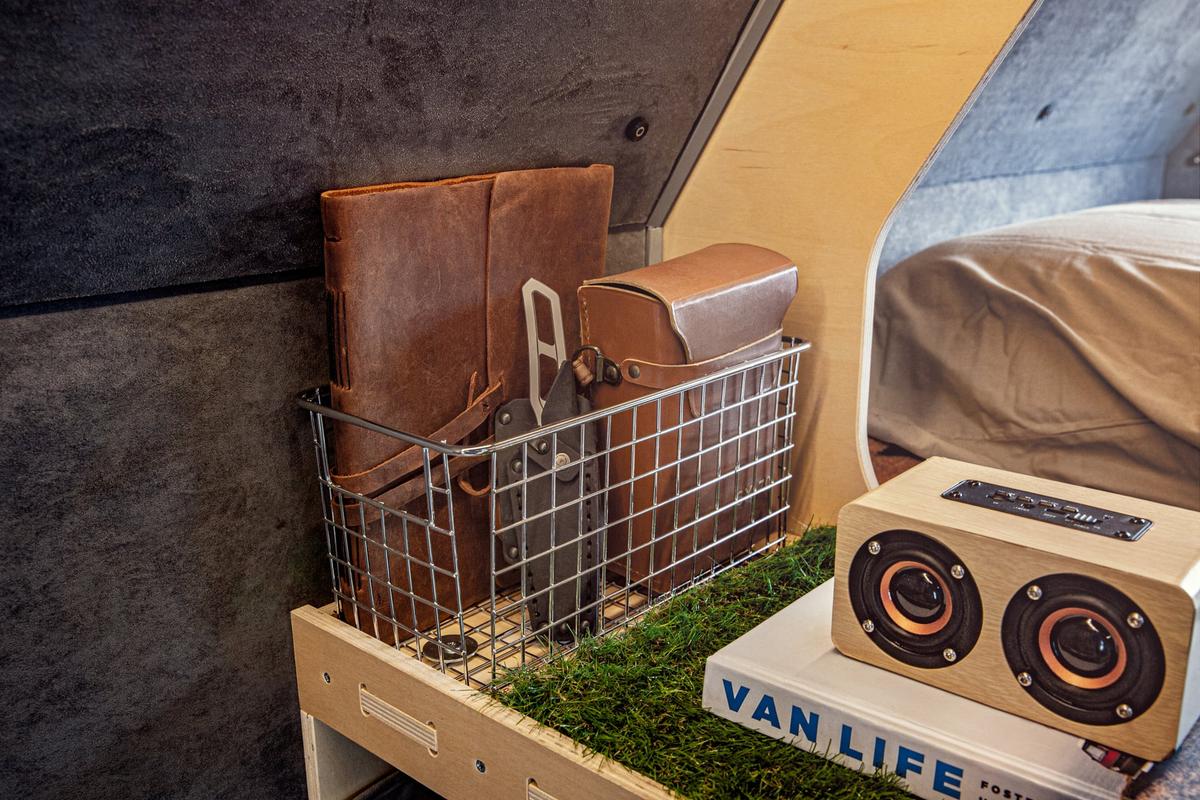Mounted wire basket inside a Kimbo 6 hard-side aluminum truck bed camper holding a hand-stitched leather notebook, a vintage Polaroid case, and a copy of a Van Life book, with a small Bluetooth speaker on the wooden shelf below.