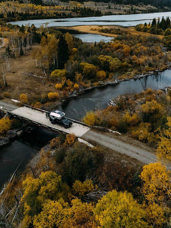 Aerial autumn view of a forested river bend in the Pacific Northwest with Kimbo owner John Smith's truck bed camper crossing a small bridge below, fall colours filling the valley.