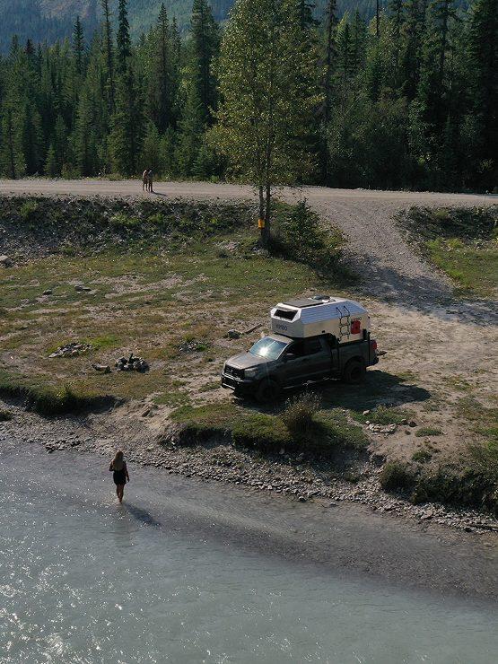 Aerial view of Kimbo owner Julie Do walking across a wide rocky river bar in the Pacific Northwest, her Kimbo truck bed camper parked at the dirt pull-off above on a forest road.