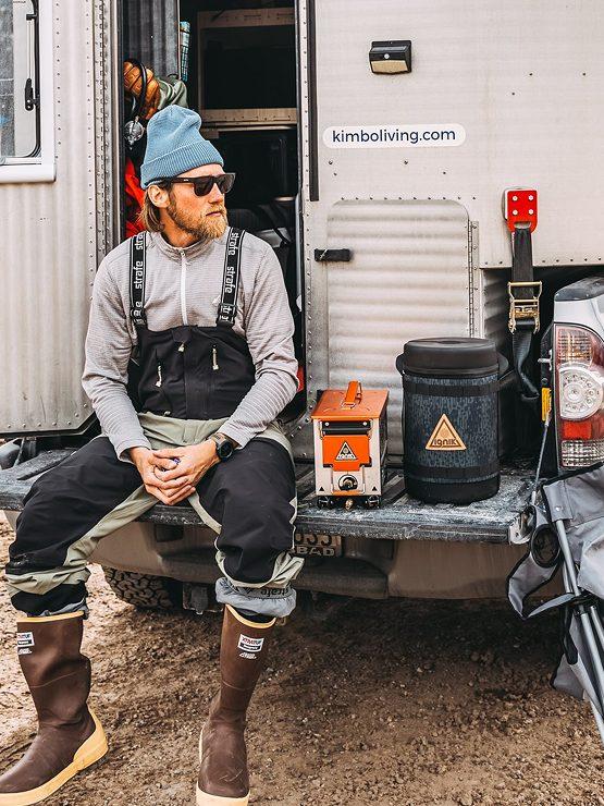 Kimbo owner Patty Klump in fishing waders sitting on the open tailgate of her white Kimbo truck bed camper, the rear cargo door swung open behind her at a forest pull-off.
