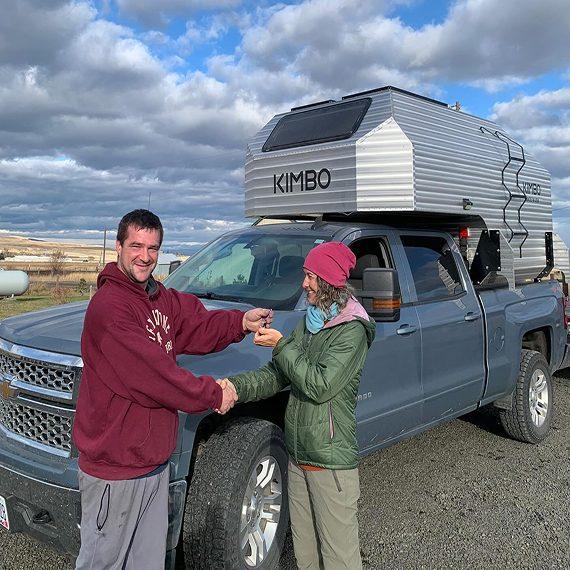 Two Kimbo owners shaking hands and exchanging keys in front of a gray Chevy Silverado with a Kimbo truck bed camper — a Bellingham factory delivery moment.