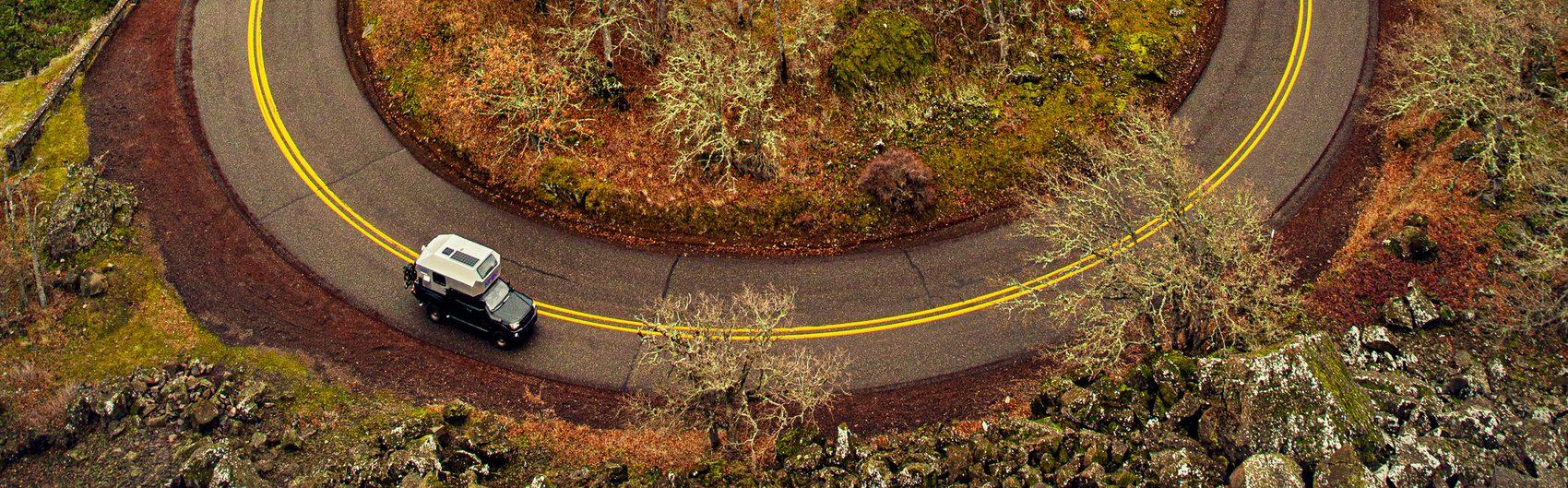 Aerial drone view of a Pacific Northwest highway switchback ringed with yellow centre-line striping, a white pickup with Kimbo hard-side aluminum truck bed camper driving the loop, autumn vegetation and basalt rock outcroppings filling the surrounding terrain.