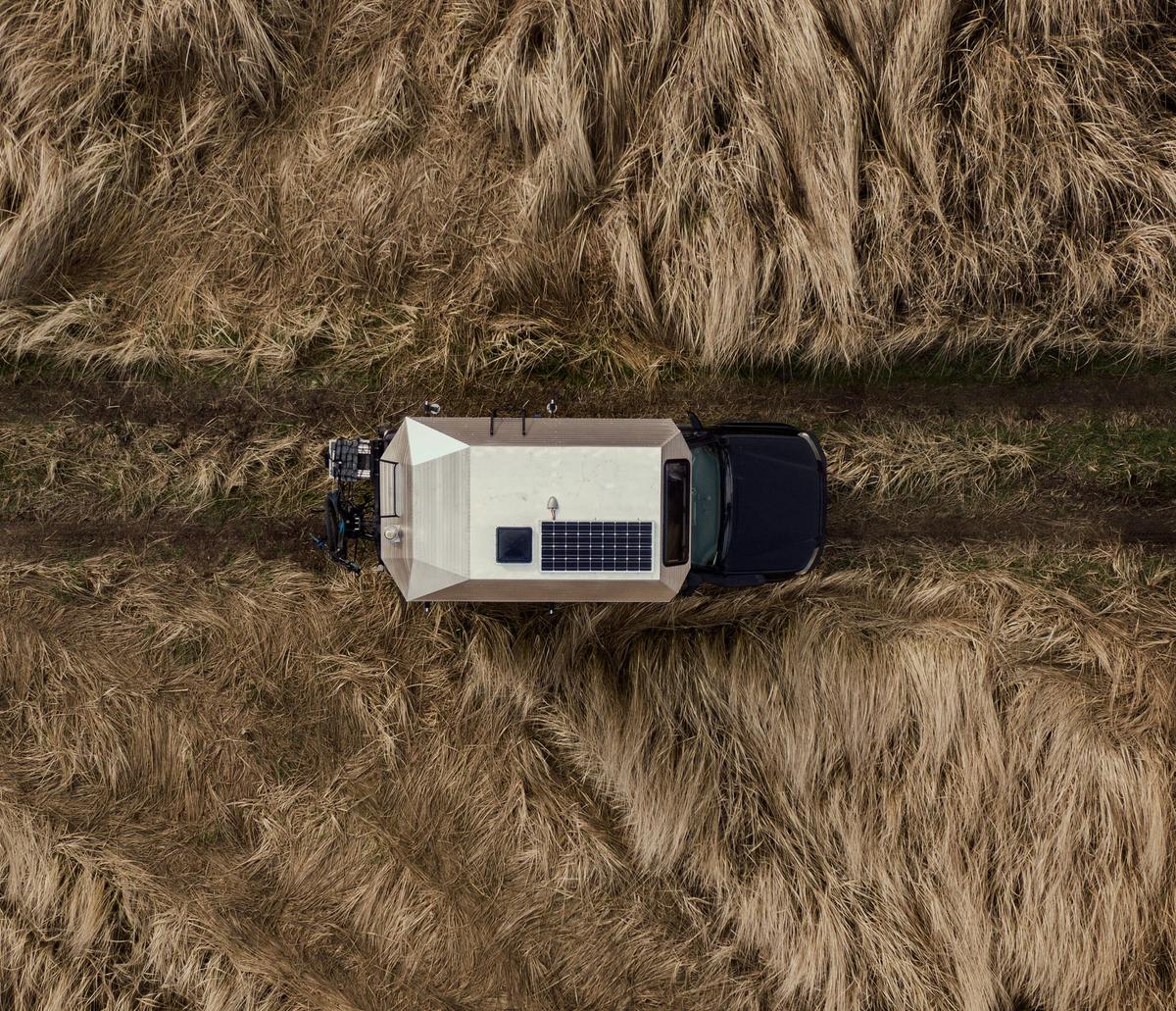 Drone-aerial top-down view of a Toyota Tacoma with hand-riveted aluminum Kimbo 6 truck bed camper on a single-track dirt road cutting through tall waving grasses, the roof-mounted solar panel visible from above.