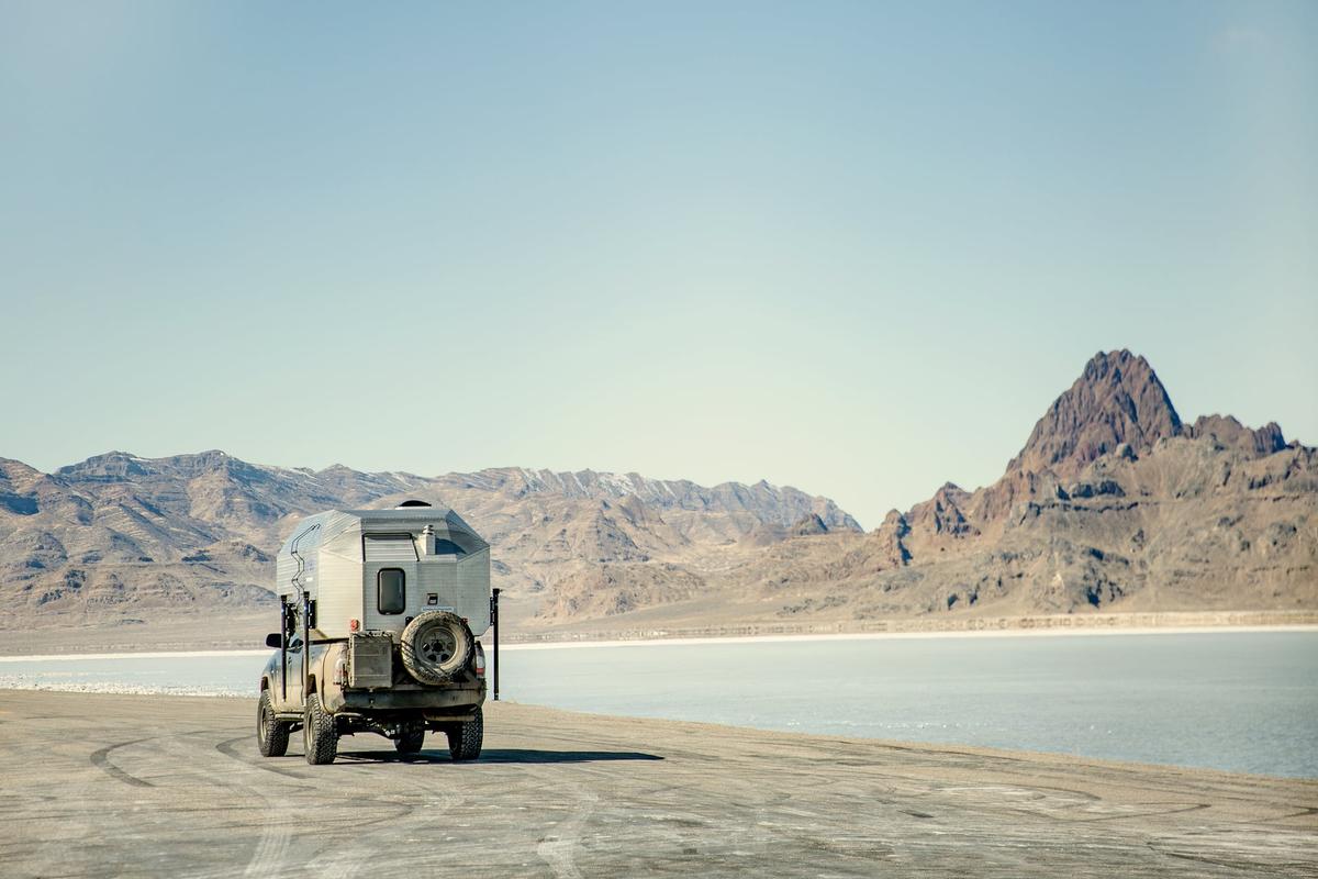 Toyota Tacoma with hand-riveted aluminum Kimbo 6 truck bed camper parked on the Bonneville Salt Flats, the white salt stretching to a row of dark rocky mountains across a lake of mirage water.