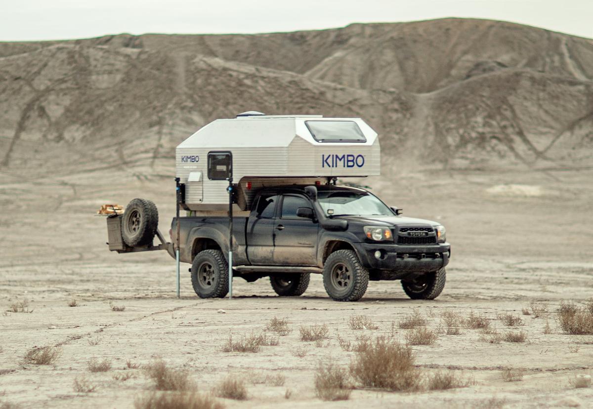 Side view of a Toyota Tacoma with hand-riveted aluminum Kimbo 6 truck bed camper raised on its four corner jacks for solo demount, parked alone on barren clay flats with eroded hills behind.