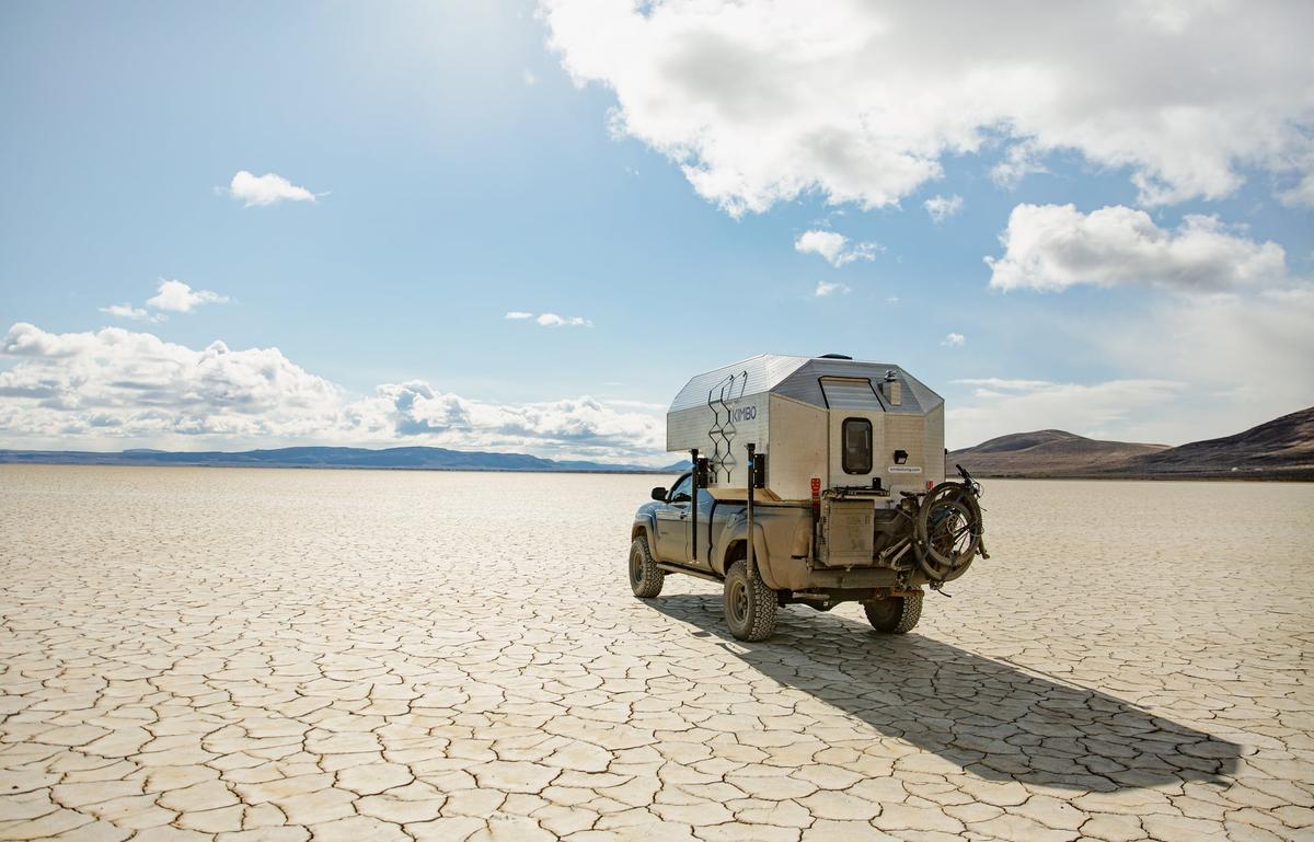 Toyota Tacoma with hand-riveted aluminum Kimbo 6 truck bed camper parked alone on a vast cracked-mud desert playa under a dramatic cloudy sky, distant mountains rimming the horizon.