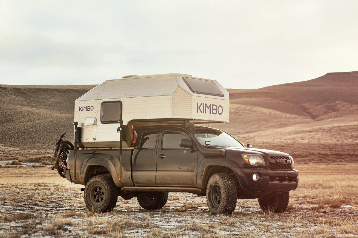 Black Toyota Tacoma with hand-riveted aluminum Kimbo 6 truck bed camper photographed side-on in dry rolling foothills at golden hour, a fat-tire bike strapped to the rear rack, KIMBO wordmark catching warm side light.