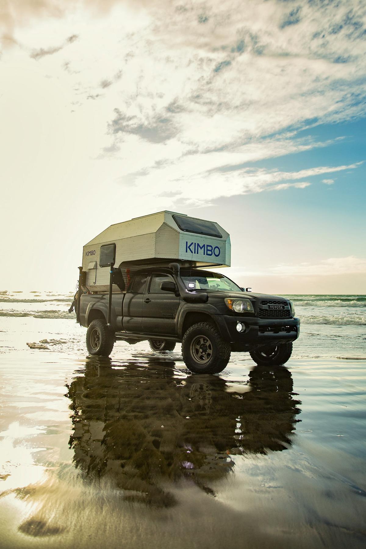 Toyota Tacoma with hand-riveted aluminum Kimbo 6 truck bed camper parked at the wet tideline of a Pacific beach at sunset, the truck silhouetted against golden water and reflective sand.