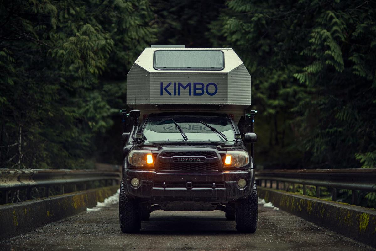 Toyota Tacoma with hand-riveted aluminum Kimbo 6 truck bed camper crossing a bridge in dense Pacific Northwest evergreen forest, headlights on against the gray daylight, KIMBO wordmark front and center.