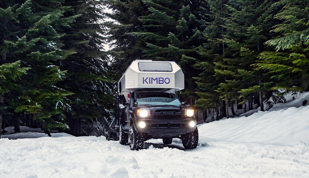 Toyota Tacoma with hand-riveted aluminum Kimbo 6 truck bed camper parked in deep Pacific Northwest snow surrounded by tall evergreens, headlights on against the wintry shade.