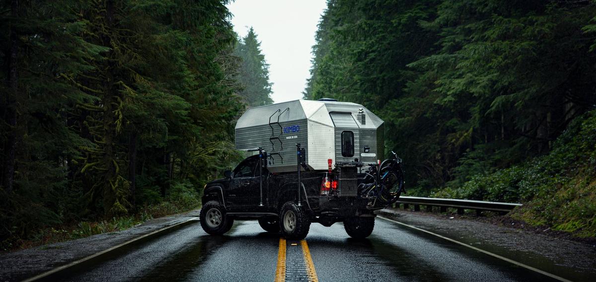 Toyota Tacoma with hand-riveted aluminum Kimbo 6 truck bed camper on a rain-slick Pacific Northwest highway lined with towering evergreens, rear three-quarter view as the truck heads into the misty distance.