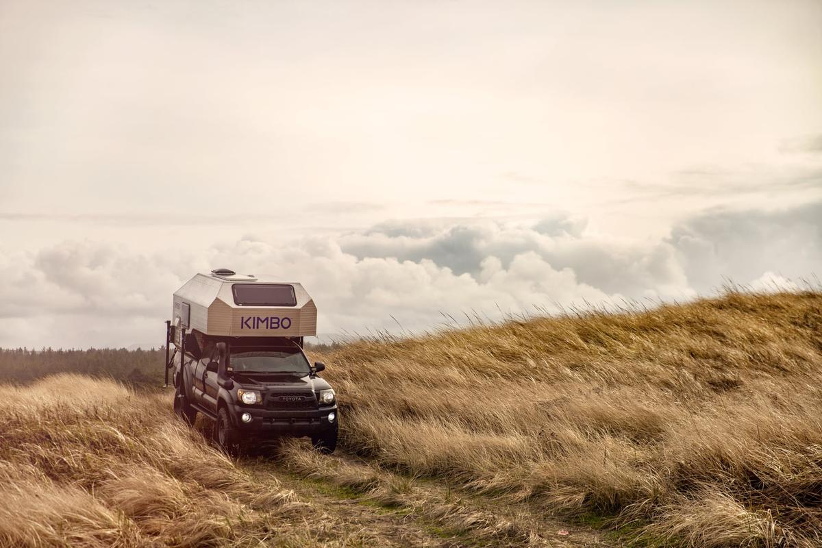 Black Toyota Tacoma with hand-riveted aluminum Kimbo 6 truck bed camper crossing a hill of golden grasses under a cloudy Pacific Northwest sky, KIMBO wordmark catching the muted afternoon light.