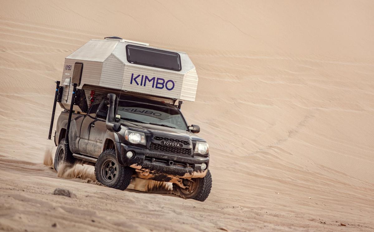 Black Toyota Tacoma with hand-riveted aluminum Kimbo truck bed camper kicking up sand as it climbs a desert dune, KIMBO wordmark prominent on the side of the camper.