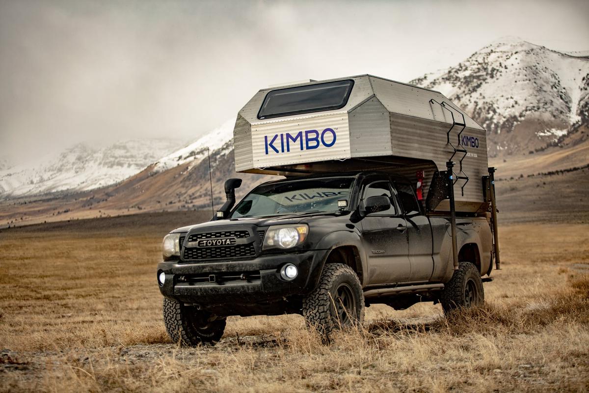 Toyota Tacoma with hand-riveted aluminum Kimbo 6 truck bed camper parked in an open meadow with a snow-covered mountain range filling the horizon behind, KIMBO wordmark prominent on the camper.