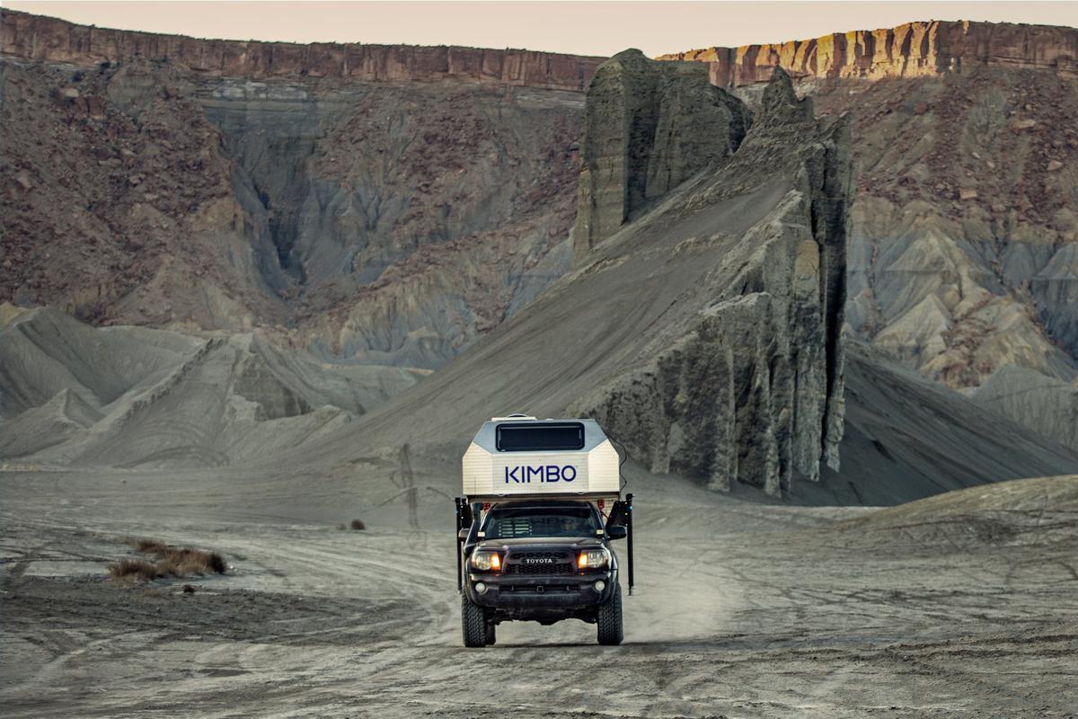 Front view of a Toyota Tacoma with hand-riveted aluminum Kimbo 6 truck bed camper driving across dramatic eroded clay badlands in Utah, headlights on, the KIMBO wordmark front and center against the spires of weathered sandstone.