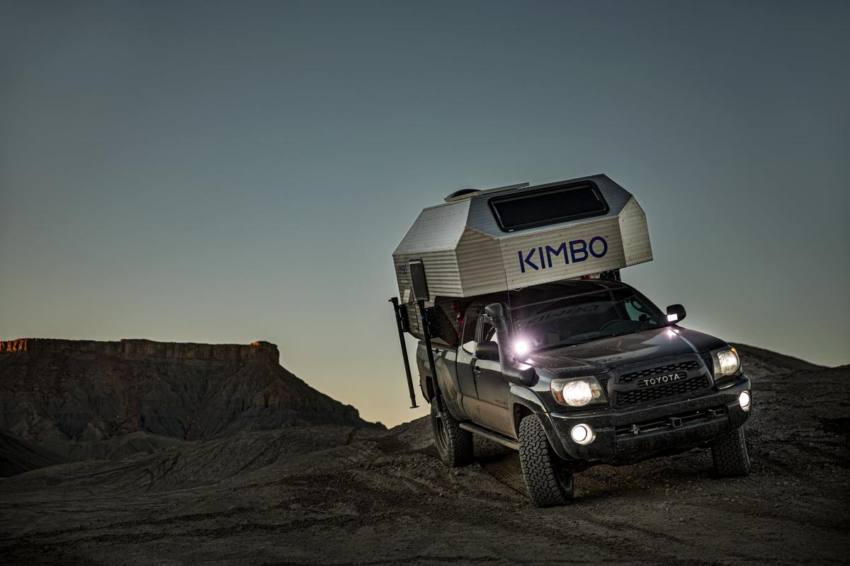 Black Toyota Tacoma with hand-riveted aluminum Kimbo 6 truck bed camper at twilight on a red rock mesa in Utah, the KIMBO wordmark catching the last of the desert sun.