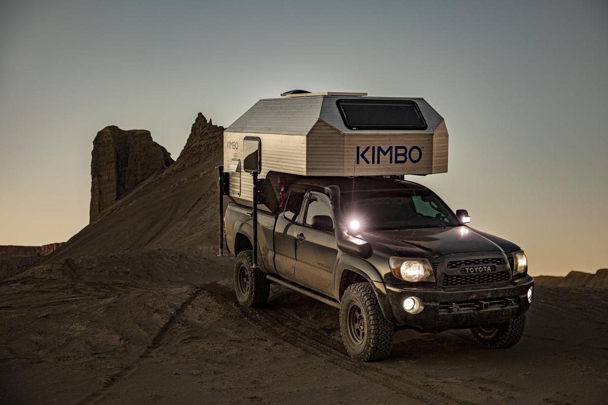 Toyota Tacoma with hand-riveted aluminum Kimbo truck bed camper at twilight beside a tall red-sandstone pinnacle in the Utah desert, KIMBO wordmark glowing against the post-sunset purple sky.
