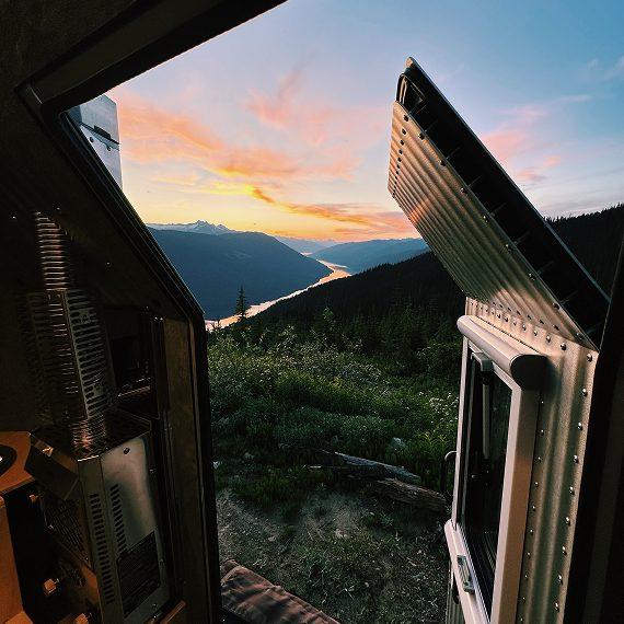 Layered alpine ridges fading into a pastel sunset, framed through the open clamshell roof of a Kimbo truck bed camper at high elevation — a Truck Camper Magazine field review viewpoint.