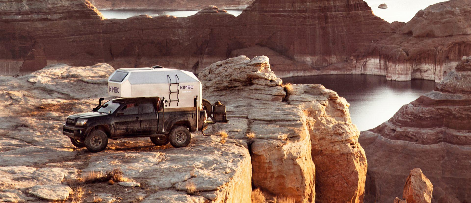 Black Toyota Tacoma with Kimbo 6 hard-side aluminum truck bed camper parked on red sandstone above a Lake Powell side canyon at golden hour, southern Utah.