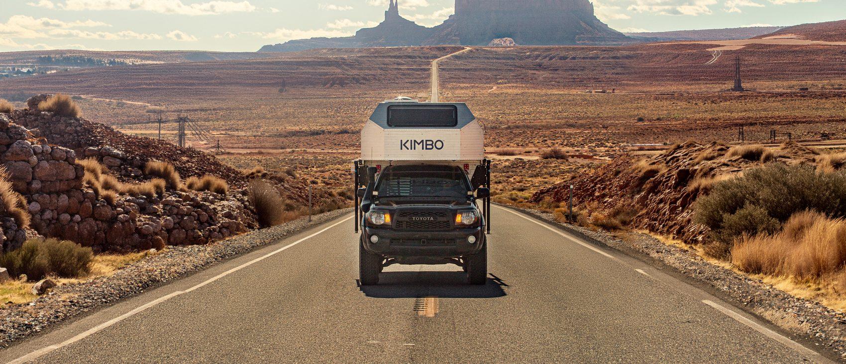 Black Toyota Tacoma with Kimbo 6 hard-side aluminum truck bed camper driving an empty desert highway toward the Monument Valley sandstone monoliths at sunset, southwest Utah.