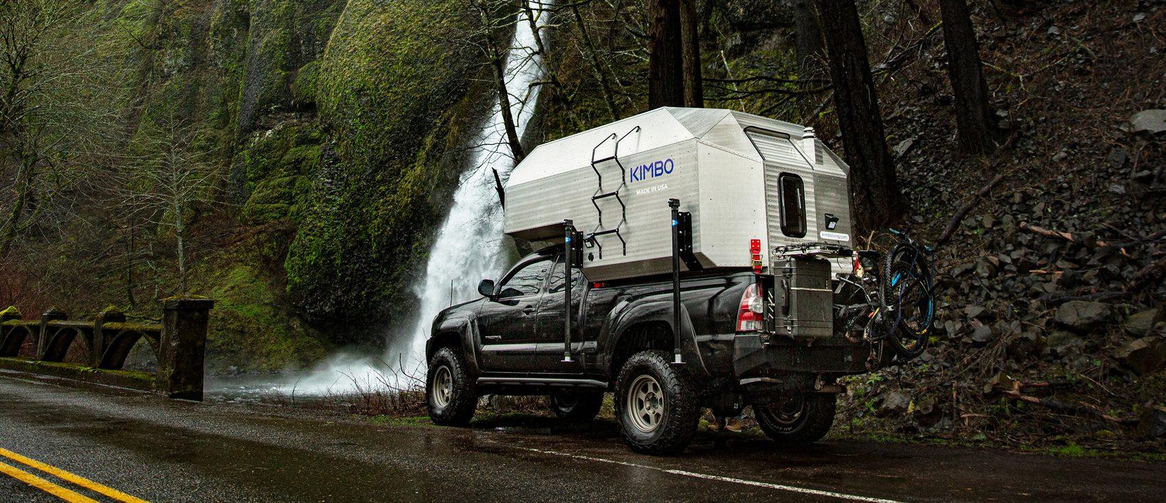Black Toyota Tacoma with Kimbo 6 hard-side aluminum truck bed camper parked beside a roaring Columbia River Gorge waterfall, a mountain bike strapped to the rear rack on a wet Pacific Northwest road.