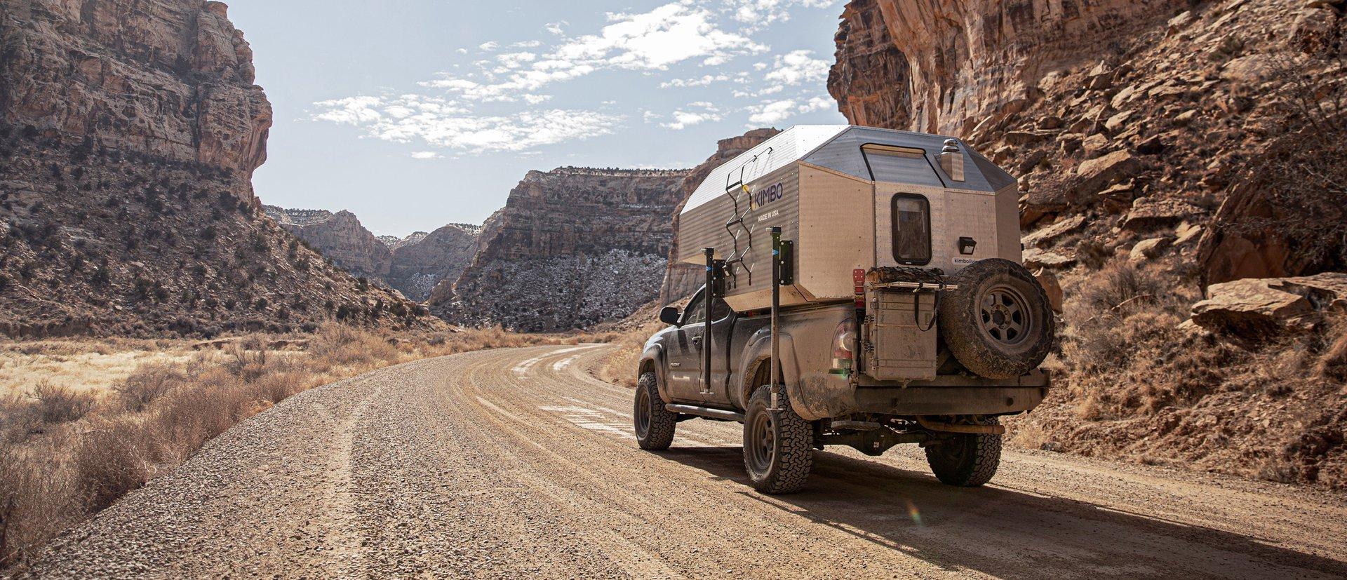 Toyota Tacoma with Kimbo 6 hand-riveted aluminum truck bed camper driving down a red-rock canyon dirt road in the Utah desert — sandstone walls on both sides, open blue sky and high clouds overhead.