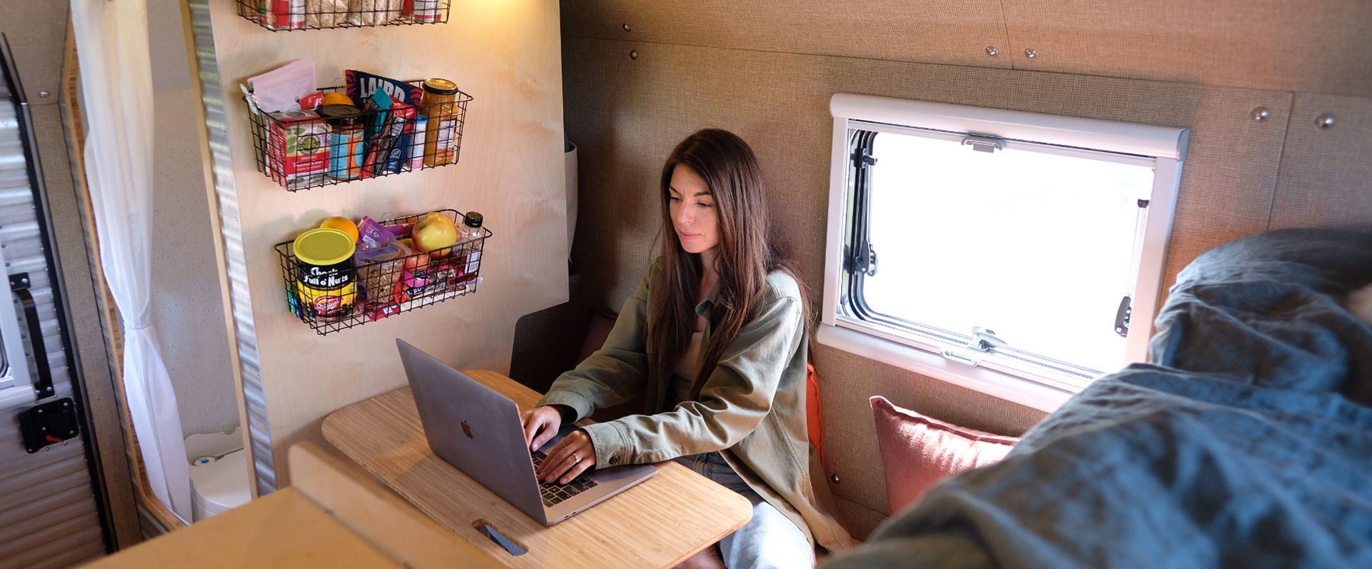 Female Kimbo 8 owner in a green long-sleeve top sitting at the bamboo dinette of her Kimbo 8 truck bed camper, working on a yellow-cased MacBook with morning light streaming through an Arctic Tern double-pane window, wire-basket pantry mounted above.