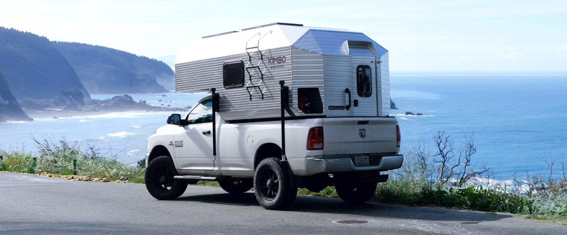 White Dodge Ram 2500 with Kimbo 8 hard-side aluminum truck bed camper parked at an Oregon coast headland pull-off, the Pacific Ocean and rugged Cape Blanco shoreline filling the background.