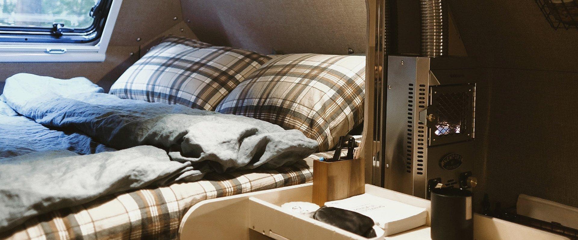 Inside the Kimbo 8 cabover — plaid pillows on a queen bed, a soft linen-look duvet pulled back, the stainless steel Dickinson propane fireplace glowing at right, and morning light coming through an Arctic Tern double-pane window above the bed.