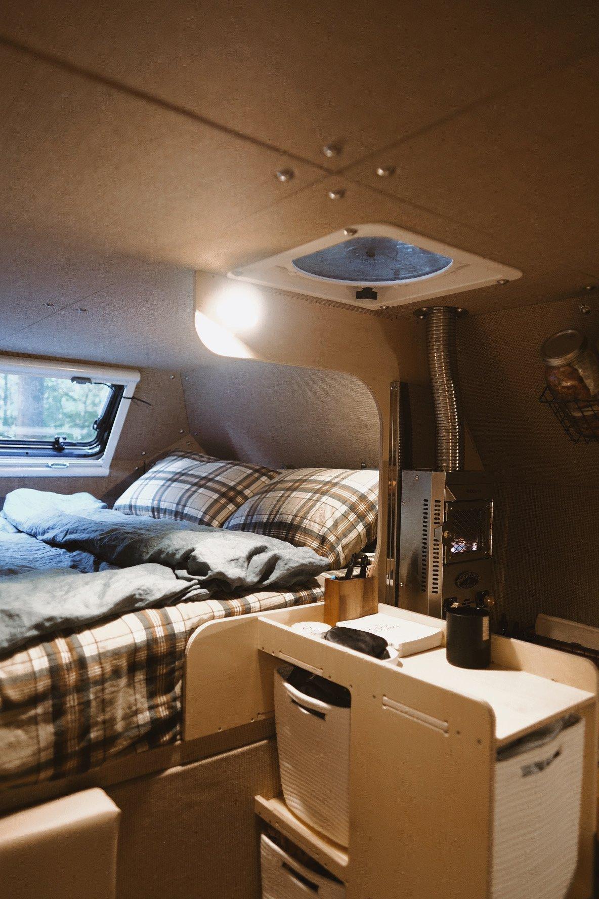 Cabover sleeping nook inside a Kimbo 8 hard-side aluminum truck bed camper — checkered plaid pillows on a queen-size memory foam mattress, twin Arctic Tern windows, MaxxAir Deluxe roof fan visible at the ceiling, and the stainless Dickinson Newport propane heater in the right corner.